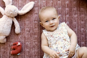 A baby wearing a floral patterned dress is lying on a textured pink blanket. The baby, perhaps destined to have one of those charming girl names starting with N, is smiling and looking up. To the baby's left, there is a stuffed rabbit and a small red stuffed animal.