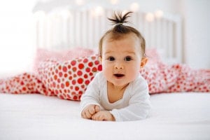 A baby girl with a small ponytail on top of her head is lying on her stomach on a bed. The baby, potentially one of those charming Girl Names That Start With D, like Daisy or Delilah, is wearing a white outfit. The bedding boasts a red and white strawberry pattern against soft lighting and a white crib in the background.