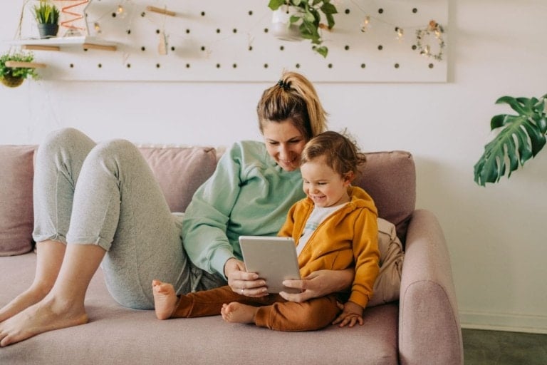 A woman and a child are sitting on a light pink couch, both smiling as they look at the tablet the woman is holding. The cozy room, adorned with string lights, wall plants, and a pegboard with small shelves, sets the perfect scene for exploring educational apps for kids.