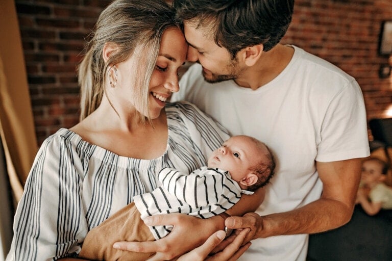 A woman and man hold a baby indoors. The woman smiles, looking at the baby, while the man leans in, touching his forehead to the woman's. Expect heartfelt moments like these during postpartum. The baby is dressed in a striped outfit. There is a brick wall in the background and a small child seated in the distance.