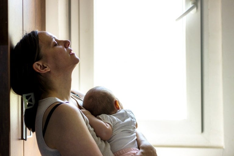 A woman sits leaning against a wall near a window, eyes closed, with a baby resting on her chest. She appears tired, possibly from birth trauma. Sunlight streams through the window, illuminating the scene. The baby is dressed in white and pink clothes.