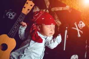A baby dressed in a white onesie wearing a red bandana is lying down with a toy microphone in one hand and a small ukulele resting beside them. The background features a black blanket with skull and crossbones patterns, perfectly capturing the essence of emo baby style.