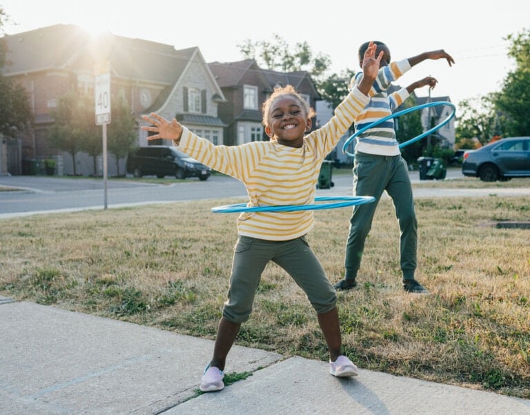 Two children are enjoying outdoor games for kids, playing with hula hoops on a residential street. The child in the foreground is smiling with arms raised while hula hooping. The child in the background is also using a hula hoop. They are both wearing striped long-sleeve shirts.
