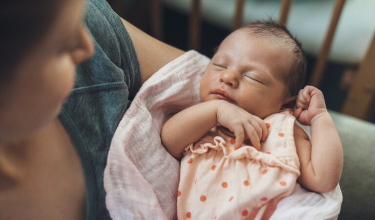 A newborn baby is sleeping peacefully, wrapped in a soft blanket with a polka dot pattern. The baby has their eyes closed and a hand near their face. An adult is holding the baby, with part of their shoulder visible in the frame—a serene moment that dispels common baby sleep myths.