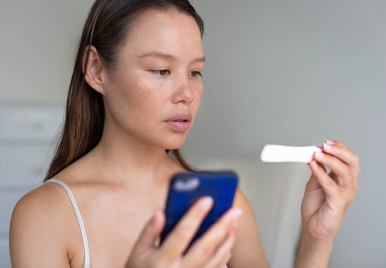 A person with long brown hair is looking intently at a pregnancy test in their right hand while holding a smartphone in their left hand, perhaps because you're trying to conceive. The person is wearing a light-colored tank top and standing indoors in a softly lit environment.