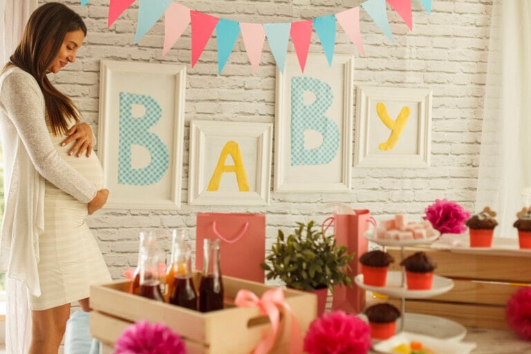 A pregnant woman in a white dress stands by a decorated table at a baby shower. The table is adorned with cupcakes, drinks, and flowers, surrounded by beautifully wrapped gifts. On the wall behind her, buntings and framed letters spell "BABY" in yellow and blue.