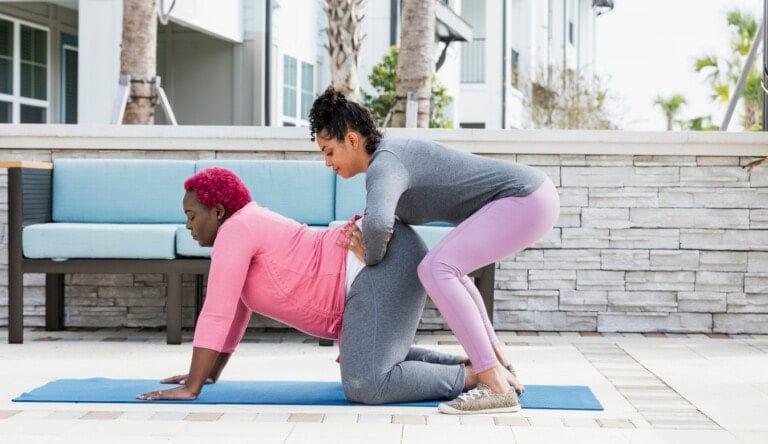 A person with pink hair in a pink shirt and gray leggings is on all fours on a blue yoga mat. Another person in gray athletic wear and sneakers is assisting by placing hands on their back, applying pressure points for labor. They are outdoors, near a stone wall and blue cushioned bench.
