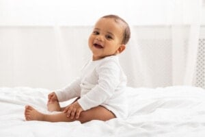 A baby named Liam, dressed in a white onesie, sits on a bed while smiling at the camera. The background is a softly lit room with white curtains.
