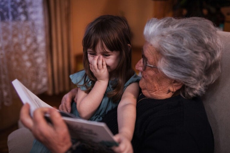 An elderly woman with gray hair and glasses sits on a chair holding a book about gratitude for kids, while a young girl with long brown hair and a blue dress sits on her lap, covering her nose and mouth with her right hand. They appear to be reading together.