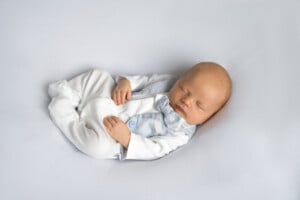 A sleeping baby, dressed in a white and light blue outfit with a bow tie, lies on a soft white surface. The baby is positioned on their back with hands gently resting on their chest, embodying the simplicity of timeless short boy names.