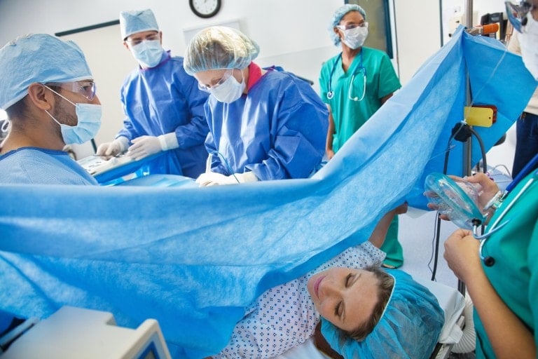 A medical team, clad in surgical attire and masks, is meticulously performing a procedure in an operating room. Behind a blue surgical drape, the patient—awake and wearing a surgical cap—lies on the table. Various medical equipment stands ready, as this might also be where c-sections are conducted.