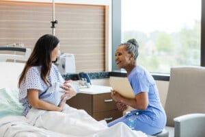 A woman holding a newborn baby sits on a hospital bed, engaging in conversation with a nurse. The nurse, in blue scrubs and holding a clipboard, explains the importance of uterine massage after birth. The room is well-lit with natural light from a window.