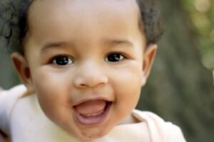 A close-up of a smiling baby with curly hair, wearing a yellow and white striped shirt. The baby, likely to be one of those charming n boy names like Noah or Nathan, is looking directly at the camera with an open mouth and bright eyes. The background is blurred and appears to be outdoors.