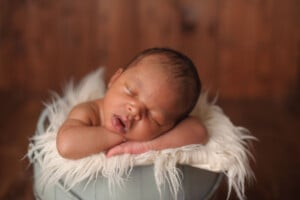 A sleeping baby rests on a fluffy white cushion inside a light blue bucket. The baby's head is tilted forward, and arms are folded under the head. The background is a warm-toned wooden surface, giving a cozy atmosphere—perfect for parents considering boy names that start with O.