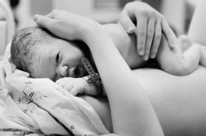 A black-and-white photo shows a newborn baby lying against a person's chest after pushing during childbirth. The baby's eyes are open and they have a hospital band on their wrist. An adult's hand is gently cradling the baby’s head and back.