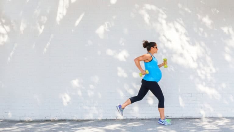 A pregnant woman in a blue tank top and black leggings is running while pregnant, holding yellow dumbbells. She moves gracefully past a white brick wall, with dappled sunlight filtering through the leaves, embodying exercise for pregnant women.