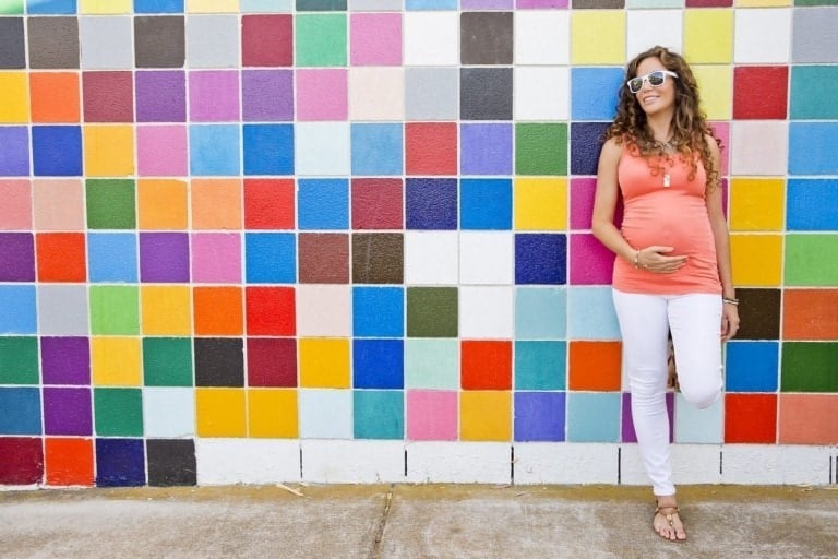 A person wearing a pink top, white pants, and white sunglasses is leaning against a colorful tiled wall, smiling and touching their stomach. This stylish look shows how you can save money while building a chic maternity wardrobe.
