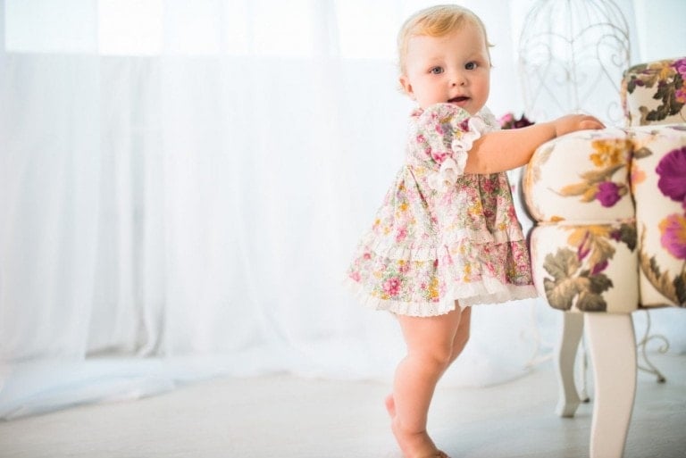A baby with blonde hair is standing and holding onto a floral patterned chair, reaching an exciting developmental milestone. Wearing a floral dress with ruffles, the baby fits perfectly in the light and airy room adorned with sheer white curtains.