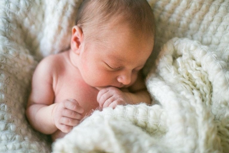 A newborn baby, symbolizing the miracle of birth, is sleeping wrapped in a soft, white knitted blanket. With eyes closed and hands gently curled near the face, the background consists of the same textured blanket.