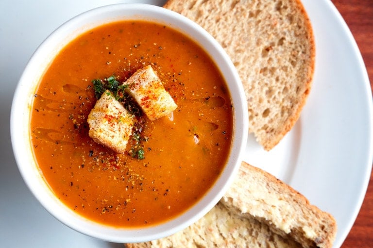 A white bowl filled with tomato soup, garnished with croutons and herbs, is placed on a white plate. Two slices of whole wheat bread are on the plate beside the bowl. The meal, inspired by batch recipes for simplicity, is set on a wooden table.