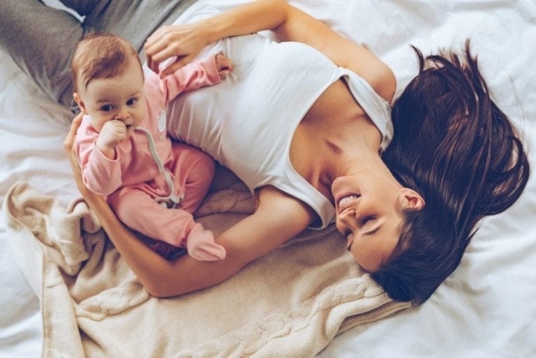 A woman with long brown hair is lying on a bed, smiling. She is wearing a white tank top and gray pants. Next to her, a baby in a pink onesie is sucking on their thumb while resting against the woman's arm. They are on a beige blanket, enjoying the cozy moment together as she thinks about getting her pre-baby body back.