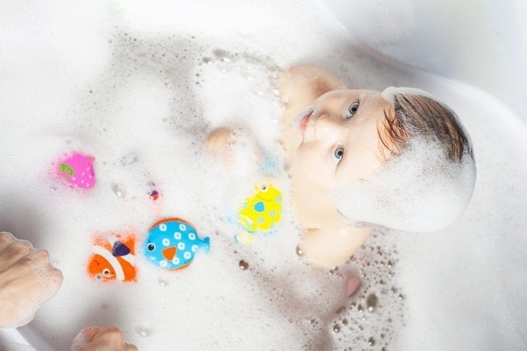 A baby with soap suds on their head is sitting in a bubble bath, surrounded by colorful floating toddler bath toys shaped like fish and a strawberry. Two hands are visible at the edge of the bathtub.