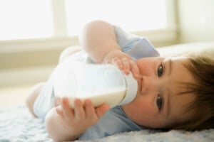 A baby in a light blue outfit lies on its back on a soft surface, holding and drinking from a bottle filled with milk. The baby has brown hair and is looking at the camera. The background includes blurred windows with bright light coming through, creating a serene scene perfect for this moment.