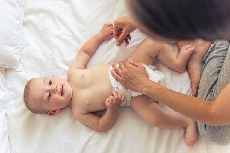 A baby is lying on a white bed while a person, potentially the parent, is changing the baby's diaper using some clever mom hacks. The baby is looking up and smiling. The person is focused on adjusting the diaper. The setting appears calm and relaxed.