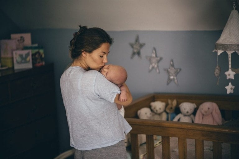 A woman kisses a baby on the head while standing in a nursery. The nursery has blue walls with star decorations and a wooden crib filled with plush toys, illustrating why some parents prefer cribs to bassinets. A chest of drawers with various items on top is visible in the background.