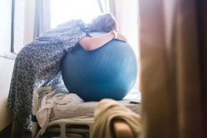 A person wearing a hospital gown leans forward onto a large blue exercise ball placed on a hospital bed, seeking natural pain relief. Sunlight streams through a window on the left, partially illuminating the scene. The room includes curtains and a chair in the background.