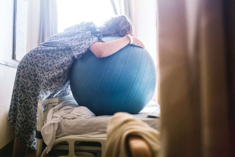 A person wearing a hospital gown leans forward onto a large blue exercise ball placed on a hospital bed, seeking natural pain relief. Sunlight streams through a window on the left, partially illuminating the scene. The room includes curtains and a chair in the background.