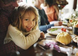 A little girl with long hair smiles while sitting at a dining table. The table is set with plates of food, a small pumpkin, and glasses of wine. A person next to the child is partially visible.