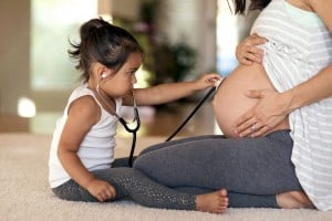 A young girl uses a stethoscope to listen to the baby bump of a pregnant woman who is kneeling on the floor, perhaps hoping to uncover some old wives tales about baby's gender. The girl is dressed in a white tank top and leggings, and the woman is wearing a striped shirt and gray leggings. They are indoors.