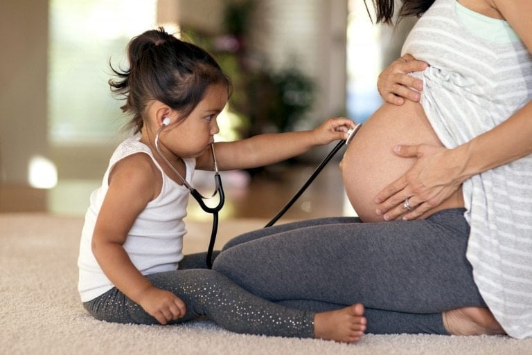 A young girl uses a stethoscope to listen to the baby bump of a pregnant woman who is kneeling on the floor, perhaps hoping to uncover some old wives tales about baby's gender. The girl is dressed in a white tank top and leggings, and the woman is wearing a striped shirt and gray leggings. They are indoors.