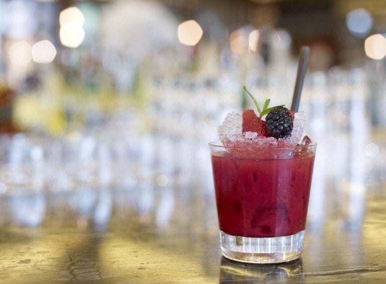 A glass of red mocktail is placed on a reflective surface. The drink is garnished with ice, a blackberry, a berry slice, and a green leaf, with a stirrer in it. The background is blurred, showcasing a bar setting. Perfect for New Year’s Mocktails Recipes!