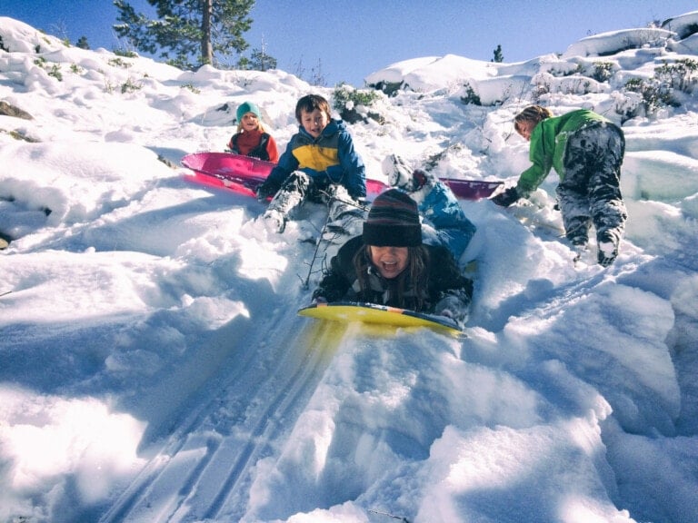 Four children are enjoying a winter playdate in the snow on a sunny day. Two kids in the foreground are riding brightly colored sleds down a snowy slope. Two others stand behind, one in a green sled and another preparing to slide. Snow-covered surroundings and trees create perfect winter playdate ideas.