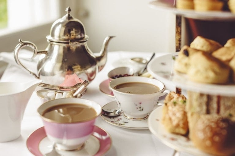 A table set for an elegant baby shower tea party, featuring a silver teapot, two teacups filled with tea, small plates, a creamer, and a three-tiered stand with assorted scones and pastries. The scene is well-lit with natural light.
