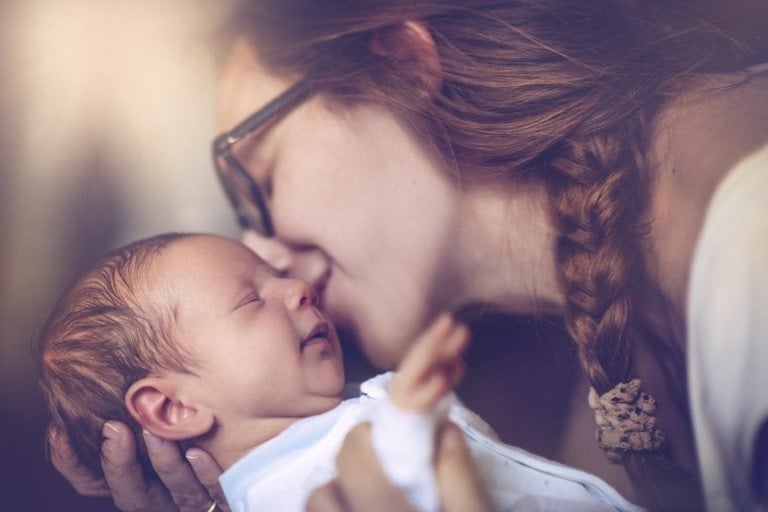 A woman with braided hair, likely navigating her postpartum recovery, is holding a newborn baby close to her face. She appears to be giving the baby a gentle kiss on the forehead. The baby is wearing a white outfit and seems content, with its eyes slightly open.