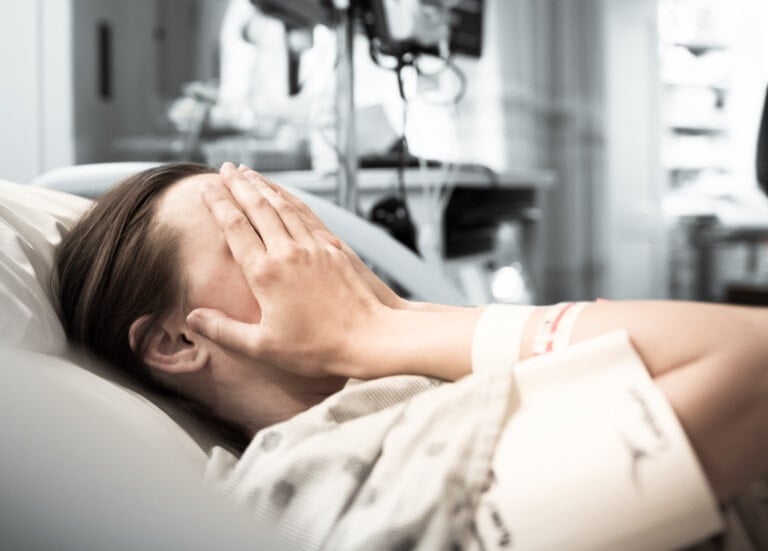 A person lying in a hospital bed covers their face with both hands. Their hospital gown and medical wristband are visible, hinting at recent birth trauma. Medical equipment and a blurred background suggest a hospital setting. The lighting is soft, creating a calm but serious atmosphere.