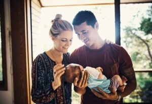 A man and a new mom are indoors near a window, both smiling and looking at the baby that the man is holding. The woman has her hand on the baby's head. The scene is lit by natural light coming through the window, with trees visible outside.