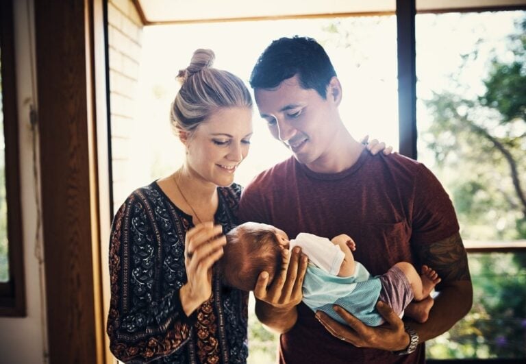 A man and a new mom are indoors near a window, both smiling and looking at the baby that the man is holding. The woman has her hand on the baby's head. The scene is lit by natural light coming through the window, with trees visible outside.