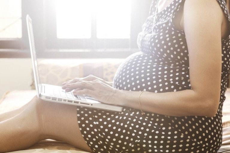 A pregnant person wearing a polka dot dress is sitting on a bed, using a laptop to explore baby registry must-haves. The bright light from the window behind them illuminates the scene, focusing on their torso and hands as they navigate through essential items for their upcoming arrival.