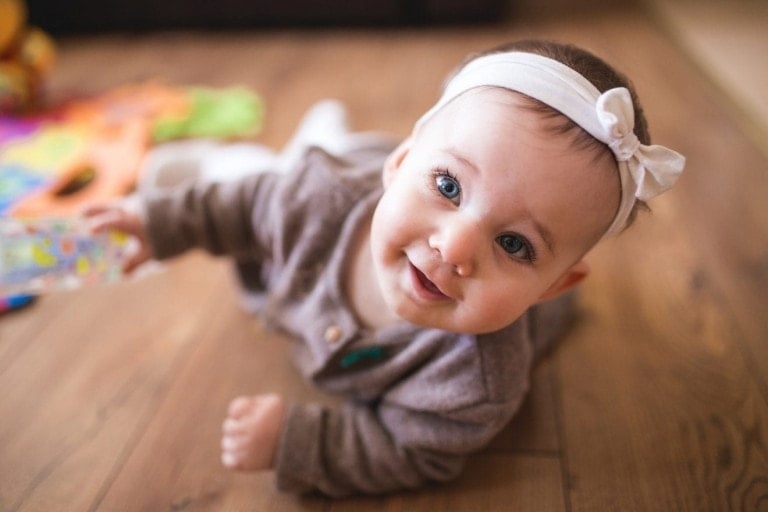 A baby wearing a brown outfit and a white headband with a bow is lying on a wooden floor. The baby is looking up and smiling, surrounded by toys in the blurred background, subtly hinting at the different ways children learn through play.