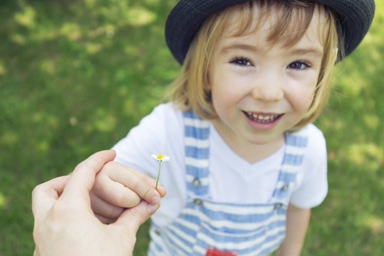 A smiling child with shoulder-length blonde hair wears a black hat, a white shirt, and striped overalls. They hold a small daisy flower graciously presented by an adult's hand, exemplifying their good manners. The background features green grass out of focus.