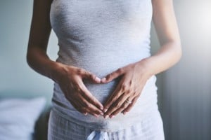 A person wearing a gray shirt and gray and white plaid pants is shown with hands placed over their stomach in a heart shape, showcasing first trimester must-haves. The setting appears to be indoors with soft light, with part of a bed visible in the background.