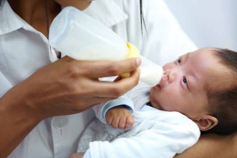 A person in a white shirt is feeding a baby with a bottle. The baby, who is wearing a blue and white outfit, is looking up at the bottle while being held. The background is gently blurred and out of focus, creating an intimate scene that contrasts with serious questions like what causes a cleft lip or palate.