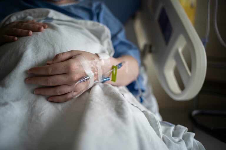 A close-up view of a person's hand resting on a blanket in a hospital bed. The hand has an IV inserted, secure with tape, highlighting the subtle difference between hep lock vs saline lock. The person is wearing a blue hospital gown, and the scene takes place within a hospital room.