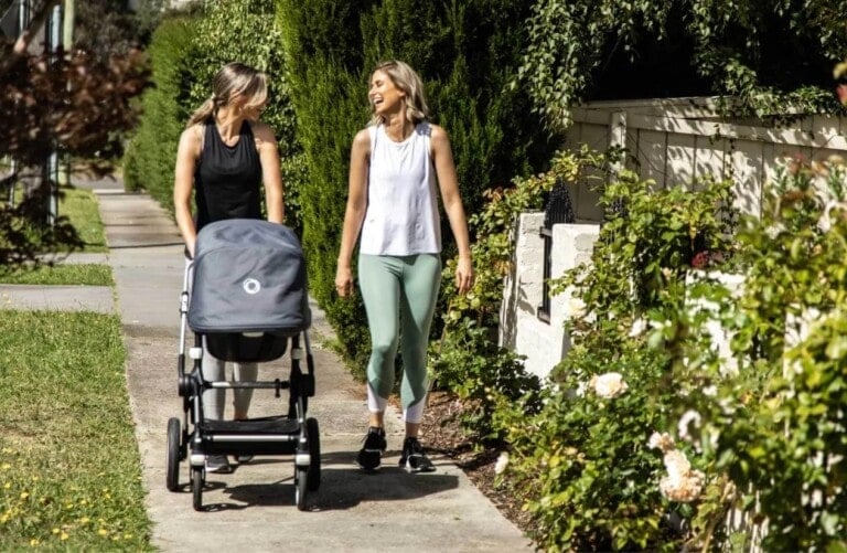 Two women are walking on a sidewalk, with one pushing a stroller. Both are casually dressed in athletic clothing and appear to be engaged in conversation, smiling. There is greenery and blooming flowers along the path—a perfect setting to boost your postpartum recovery.
