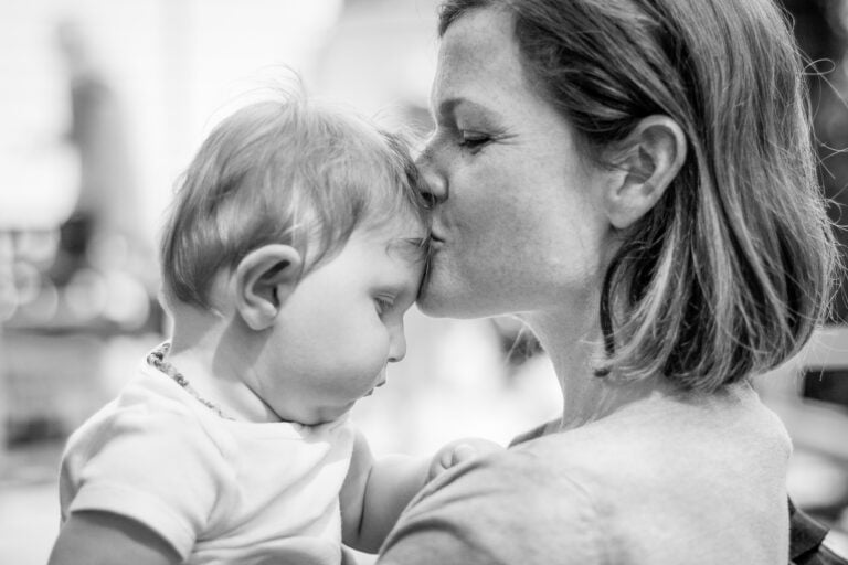 A black and white photo captures a woman gently kissing the forehead of a baby she is holding close. The baby, possibly a disabled child, rests his head against the woman's cheek, creating a tender and affectionate parenting moment. The background is blurred.