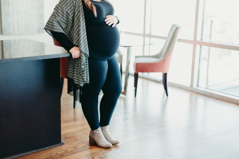 A person stands indoors next to a kitchen counter, wearing a black outfit and shawl, light brown ankle boots, and resting their hand on a visibly pregnant belly. There is a table and two chairs in the background, near a window—the perfect setting for learning how to style leggings during pregnancy.